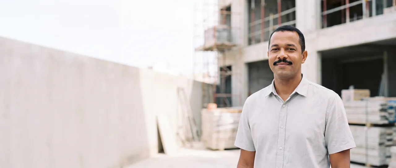 Comprador de construtora, de camisa cinza-clara, sorri satisfeito no canteiro de obras, com a estrutura de concreto e os andaimes desfocados ao fundo, de dia.