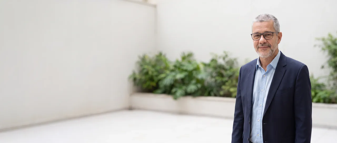 Diretor comercial de blazer azul-marinho e óculos sorri, tranquilo, no pátio interno claro da empresa, com canteiro de plantas e parede lisa desfocados, de dia.