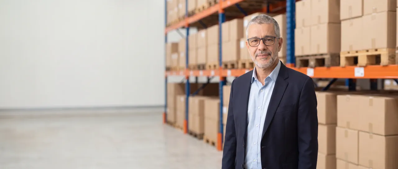 A distributor's director in a navy blazer and glasses smiles calmly in a bright distribution center aisle, with pallet racks out of focus behind him.