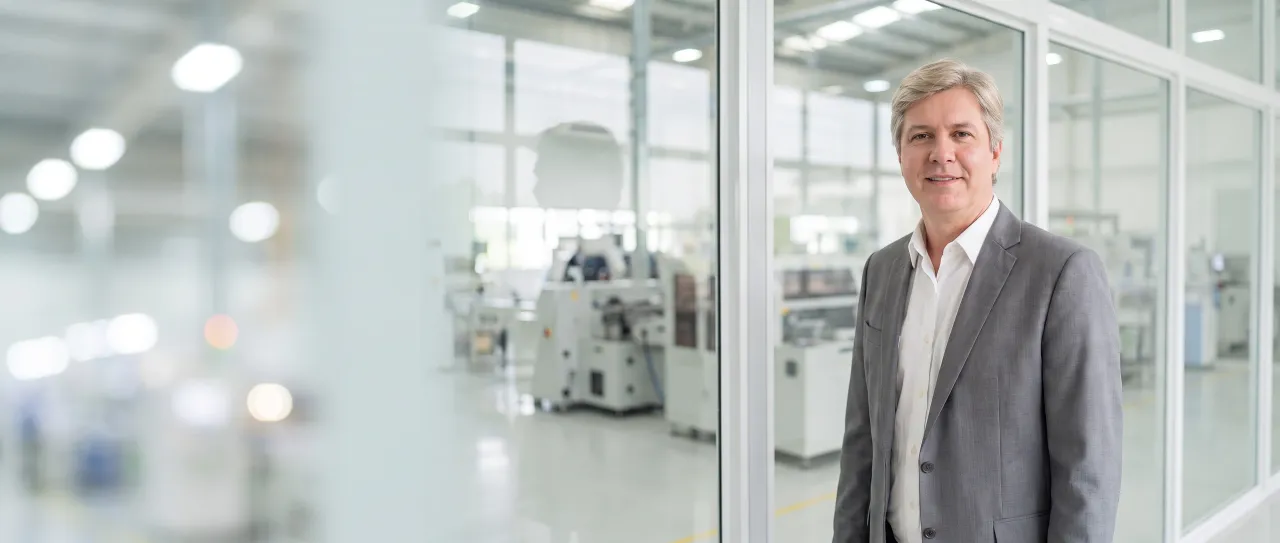 A manufacturer's director in a grey suit with no tie smiles calmly in a glass-walled office above the factory floor, with the production line out of focus.
