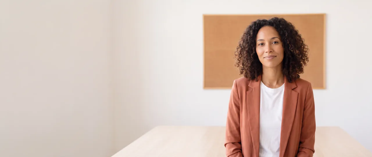 Diretora de marketing de blazer terracota sorri, tranquila, numa sala de reunião clara, com painel de cortiça vazio e mesa clara desfocados ao fundo, de dia.