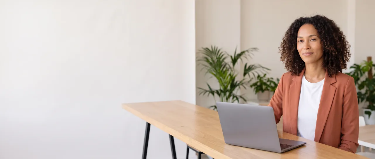 Diretora de marketing de blazer terracota sorri, tranquila, junto à mesa alta da área de convivência da empresa, com um laptop aberto e plantas desfocadas.