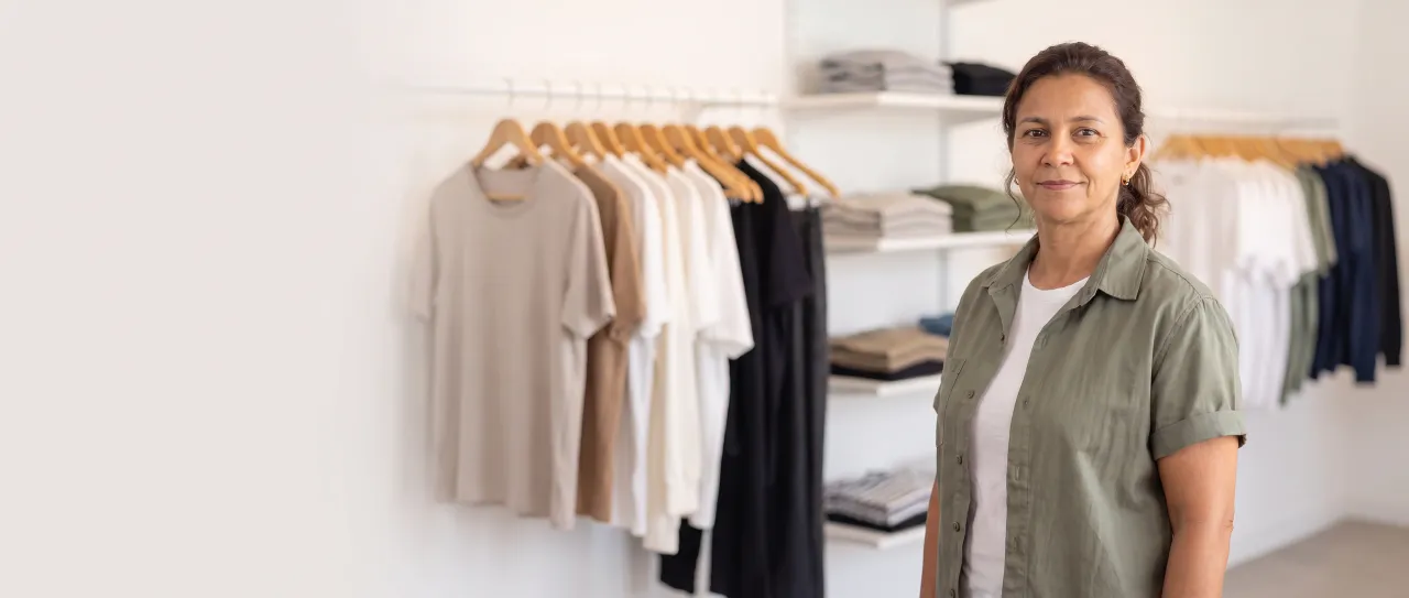 Lojista de uma loja multimarca de roupas sorri satisfeita ao lado das araras, com peças lisas penduradas e prateleiras desfocadas ao fundo, em ambiente claro.